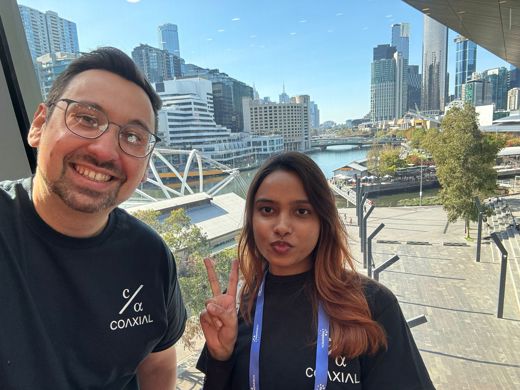 Two people wearing matching branded Coaxial AI t-shirts in front of a scenic view of Sydney.