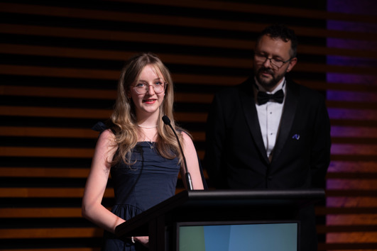 A young woman in a dark blue dress stands in a spotlight at a lectern, while a man in a black suit and bowtie looks on from the side.